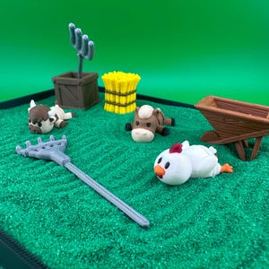 May include: A miniature farm scene with a rake, wheelbarrow, hay bale, and various farm animals including a cow, horse, and chicken. The scene is set on a green surface, with a brown wooden box and a cactus.