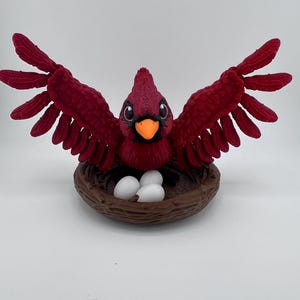 May include: A vibrant red cardinal bird figurine with outstretched wings, perched above a brown nest containing three white eggs. The bird has a black beak and eyes, and the wings are detailed with textured feathers. A decorative piece.