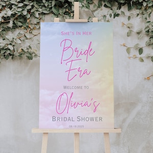 Może przedstawiać: Pastelowe tło tęczy z chmurami i tekstem "She's in her Bride Era" w różowym piśmie. Pod tekstem znajduje się "Welcome to Olivia's Bridal Shower" w różowym piśmie i data "08.18.2025" w szarym kolorze.