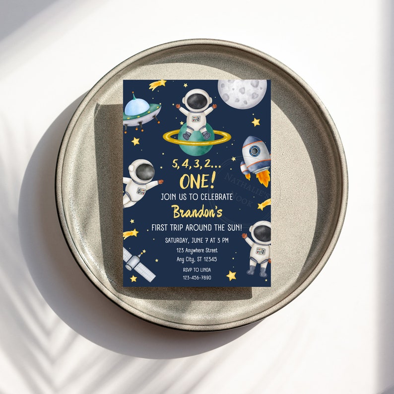 May include: A blue and yellow space-themed first birthday party invitation with a cartoon astronaut, a rocket ship, and stars. The text reads "5, 4, 3, 2... ONE! Join us to celebrate Brandon's First Trip Around the Sun! Saturday, June 7 at 3 PM 123 Anywhere Street Any City, ST 12345 RSVP to Linda 123-456-7890"