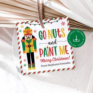 May include: A square Christmas gift tag with a nutcracker illustration. The tag features the text "GO NUTS and PAINT ME" in green and red, with "Merry Christmas!" and the name "Stephanie Gonzalez" below. The tag has a brown string.