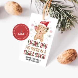 Könnte beinhalten: Ein weißes Geschenk-Etikett mit einem Lebkuchenmann mit Weihnachtsmütze, der Geschenke hält. Der Text lautet "Thank you for making me a Smart Cookie" in Grün und Rot. Verziert mit Schneeflocken und einem Lederband.