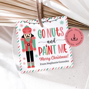 May include: A square Christmas gift tag with a nutcracker illustration. The tag features the text "GO NUTS and PAINT ME Merry Christmas!" in red and green, with a name at the bottom. The tag has a string for hanging.