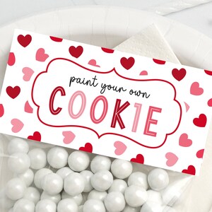 Puede incluir: Bolsas de regalo blancas con una etiqueta blanca que dice "pinta tu propia galleta" en texto negro. La etiqueta est&aacute; decorada con corazones rosas y rojos.