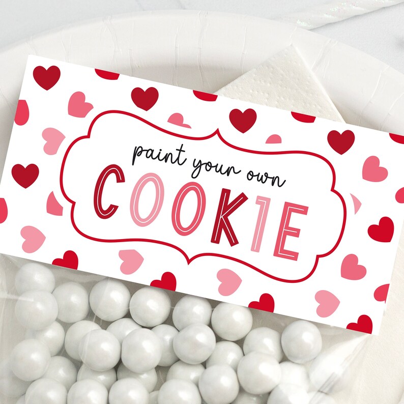 Puede incluir: Bolsas de regalo blancas con una etiqueta blanca que dice "pinta tu propia galleta" en texto negro. La etiqueta est&aacute; decorada con corazones rosas y rojos.
