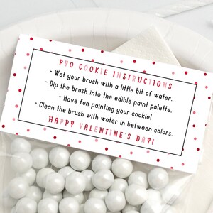 Puede incluir: Una tarjeta rosa y blanca con lunares rojos y texto negro que dice "PYO COOKIE INSTRUCTIONS - Moja tu pincel con un poco de agua. Sumerge el pincel en la paleta de pintura comestible. - &iexcl;Divi&eacute;rtete pintando tu galleta! Limpia el pincel con agua entre colores. &iexcl;FELIZ D&Iacute;A DE SAN VALENT&Iacute;N!"