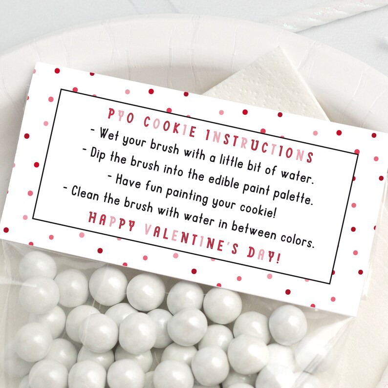 Puede incluir: Una tarjeta rosa y blanca con lunares rojos y texto negro que dice "PYO COOKIE INSTRUCTIONS - Moja tu pincel con un poco de agua. Sumerge el pincel en la paleta de pintura comestible. - &iexcl;Divi&eacute;rtete pintando tu galleta! Limpia el pincel con agua entre colores. &iexcl;FELIZ D&Iacute;A DE SAN VALENT&Iacute;N!"