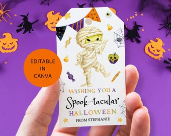 May include: A Halloween-themed gift tag featuring a cartoon mummy with the text "Wishing you a Spook-tacular Halloween". The tag includes decorative elements like candy corn, a spider web, and a jack-o'-lantern. The tag is held in a hand.