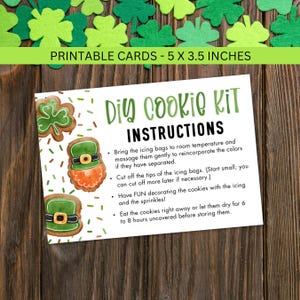 Puede incluir: Una tarjeta blanca con el texto "DIY COOKIE KIT INSTRUCTIONS" e ilustraciones de galletas decoradas. La tarjeta mide 12,7 x 8,9 cm. Decoraciones de tréboles verdes están esparcidas alrededor de la tarjeta, con más tréboles en el fondo.