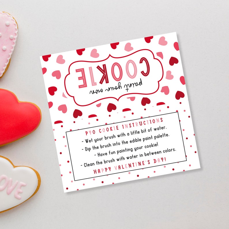 Puede incluir: Una tarjeta blanca con corazones rojos y rosas impresos en ella. La tarjeta tiene instrucciones para una actividad de "Pinta tu propia galleta". El texto dice "Instrucciones para la galleta de pintar: Moja tu pincel con un poco de agua. Sumerge el pincel en la paleta de pintura comestible. &iexcl;Divi&eacute;rtete pintando tu galleta! Limpia el pincel con agua entre colores. &iexcl;Feliz D&iacute;a de San Valent&iacute;n!"