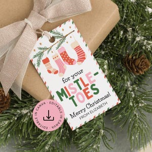 Puede incluir: Una etiqueta de regalo navideña con un paquete de papel marrón y una cinta beige. La etiqueta tiene el texto "for your MISTLE-TOES" y "Merry Christmas!" con un diseño de calcetines navideños colgantes.