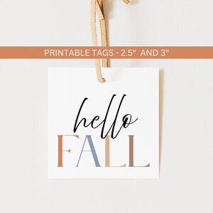 May include: A square white tag with the words "hello FALL" in black and muted orange, blue, and brown lettering. The tag is suspended by a tan string. The text "PRINTABLE TAGS - 2.5" AND 3"" is printed on a brown banner above the tag.