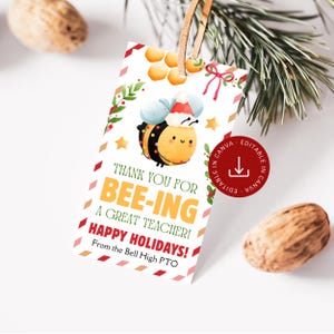 Op de afbeelding: Een wit cadeaulabel met een cartoonbij met een kerstmuts. Op het label staat "Bedankt voor het zijn van een geweldige leraar! Fijne feestdagen!" met een rode en gouden rand en een bruin touwtje.