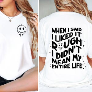 May include: A white short-sleeve t-shirt featuring a small black melting smiley face graphic on the front left chest. The back displays the black text "WHEN I SAID I LIKED IT ROUGH I DIDN'T MEAN MY ENTIRE LIFE" with a melting smiley face icon.