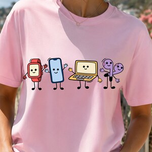 Może przedstawiać: Jasnoróżowy t-shirt z krótkim rękawem z nadrukiem graficznym przedstawiającym cztery antropomorficzne urządzenia elektroniczne. Wzór zawiera czerwony smartwatch, niebieski smartfon, żółty laptop oraz fioletowe słuchawki bezprzewodowe z buźkami.