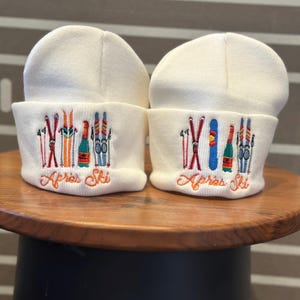 May include: Two white knit beanies with embroidered ski-themed designs and the text "Apres Ski".