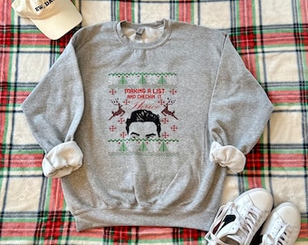 May include: A gray sweatshirt with the text "MAKING A LIST AND CHECKIN IT Twice" and a graphic of a person's face, reindeer, and Christmas trees. A beige baseball cap with "EW, DAVID" is next to the sweatshirt. White sneakers are also in the image.