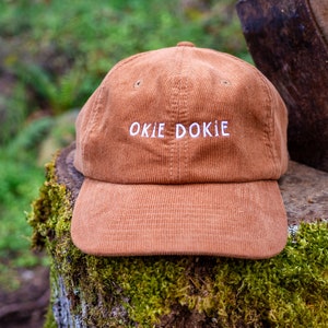 May include: A brown corduroy baseball cap with white embroidery that reads "OKIE DOKIE". The cap is sitting on a tree stump covered in moss.