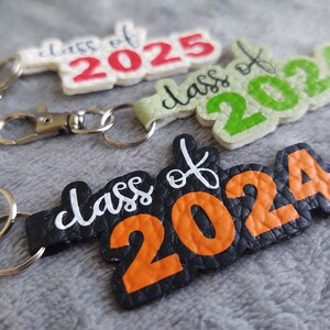 May include: Three keychains with different colored backgrounds and the text "Class of" and the year. The first keychain is white with red text, the second is green with white text, and the third is black with orange text.