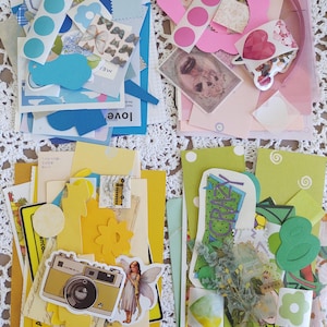 May include: A collection of paper ephemera organized into four color-themed piles: blue, pink, yellow, and green. The assortment includes die-cut shapes, stickers, vintage-style illustrations, floral patterns, and graphic prints featuring a camera and fairy.