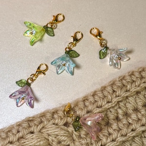 May include: Five flower-shaped charms with gold clasps and green leaf details. The flowers are in green, blue, white, purple, and pink. The charms are displayed on a beige crocheted surface.