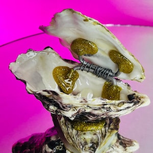 May include: A white oyster shell with a silver wire and gold glitter accents. The oyster shell is open and has a gold glitter design inside.