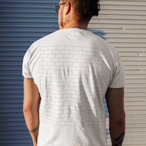 Pode incluir: Uma camiseta branca com um design de parede de tijolos preto e branco nas costas. A camiseta est&aacute; sendo usada por uma pessoa com a cabe&ccedil;a raspada e um coque.