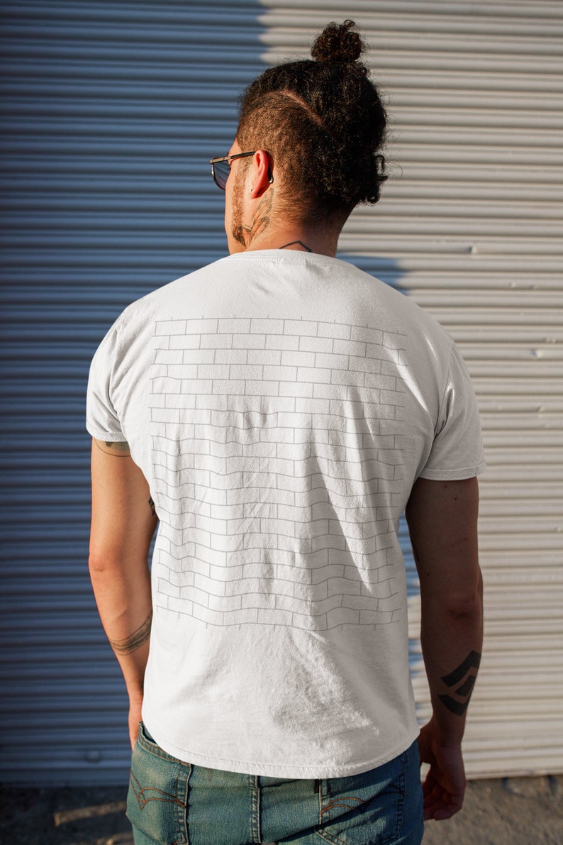 Pode incluir: Uma camiseta branca com um design de parede de tijolos preto e branco nas costas. A camiseta est&aacute; sendo usada por uma pessoa com a cabe&ccedil;a raspada e um coque.
