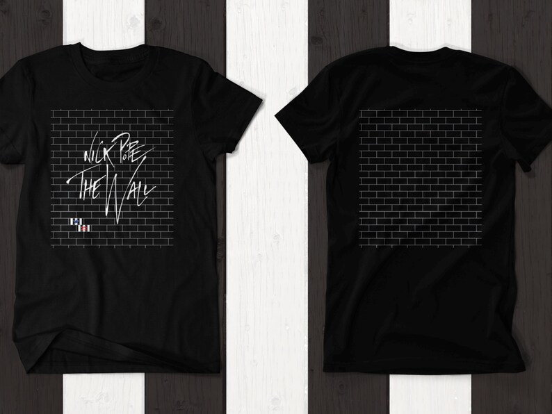 Pode incluir: Camiseta preta com um gr&aacute;fico branco de uma parede de tijolos e o texto "Nick Lowe The Wall". A parte de tr&aacute;s da camiseta apresenta um gr&aacute;fico branco de uma parede de tijolos.