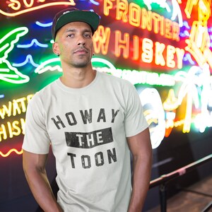 May include: A person wearing a white t-shirt with black text that reads "Howay The Toon" in a vintage style. The person is standing in front of a neon sign that says "Frontier Whiskey".