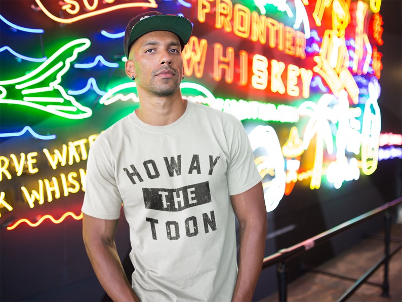 May include: A person wearing a white t-shirt with black text that reads "Howay The Toon" in a vintage style. The person is standing in front of a neon sign that says "Frontier Whiskey".