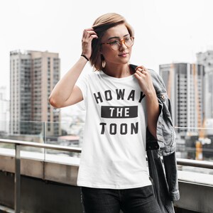 Op de afbeelding: Een wit T-shirt met zwarte tekst die "Howay the Toon" zegt. De persoon die het shirt draagt staat op een balkon met een skyline op de achtergrond.