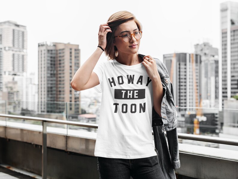 Op de afbeelding: Een wit T-shirt met zwarte tekst die "Howay the Toon" zegt. De persoon die het shirt draagt staat op een balkon met een skyline op de achtergrond.