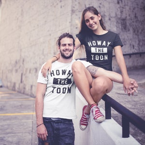 Op de afbeelding: Een stel draagt ​​bijpassende zwart-witte T-shirts met de tekst "Howay the Toon" erop gedrukt. De vrouw draagt ​​een zwart T-shirt en de man draagt ​​een wit T-shirt. Ze staan ​​voor een bakstenen muur.