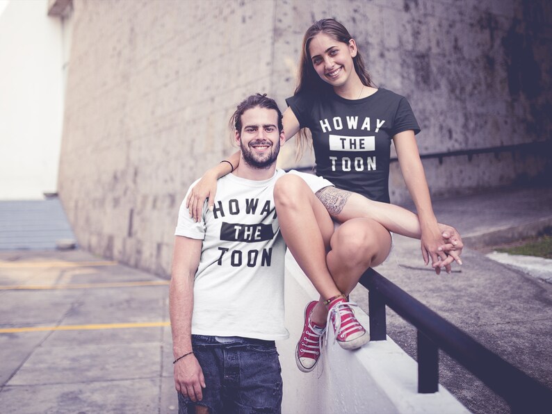 Op de afbeelding: Een stel draagt ​​bijpassende zwart-witte T-shirts met de tekst "Howay the Toon" erop gedrukt. De vrouw draagt ​​een zwart T-shirt en de man draagt ​​een wit T-shirt. Ze staan ​​voor een bakstenen muur.