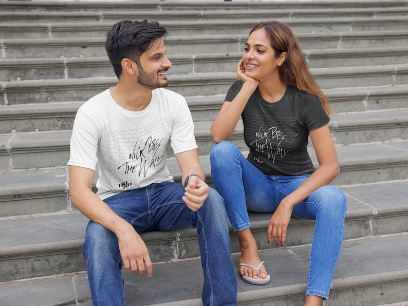 Pode incluir: Duas pessoas vestindo camisetas com um gr&aacute;fico de uma parede de tijolos e o texto "Nick Cave The Wall" nela. As pessoas est&atilde;o sentadas em um conjunto de escadas de concreto.