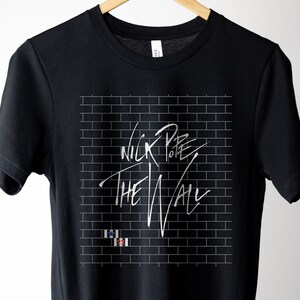 Pode incluir: Camiseta preta com um design de parede de tijolo branco e o texto "Nick Pope The Wall" em escrita branca.