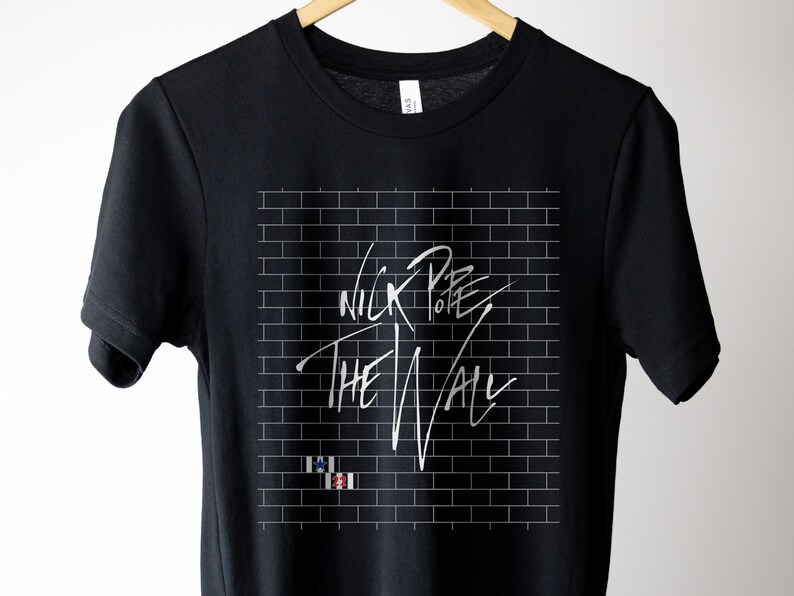 Pode incluir: Camiseta preta com um design de parede de tijolo branco e o texto "Nick Pope The Wall" em escrita branca.
