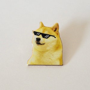May include: A yellow dog-like character wearing pixelated black sunglasses. The image is a cut-out sticker or pin, with a white background. The dog has a smug expression, and the sunglasses add a humorous touch. The design is simple and eye-catching.