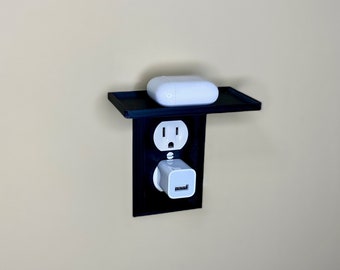 Puede incluir: Una estantería negra montada en la pared con una toma de corriente blanca y un cargador USB blanco. Una plataforma de carga inalámbrica blanca está en la estantería.