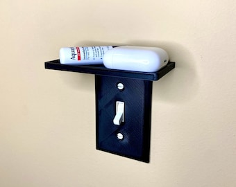 Puede incluir: Una tapa de interruptor de luz negra impresa en 3D con un pequeño estante adjunto. El estante sostiene un tubo de pomada Aquaphor y una caja blanca para auriculares inalámbricos.