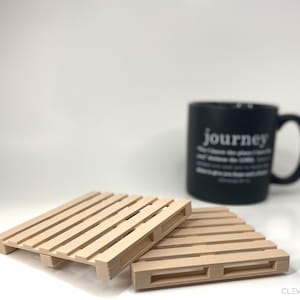 May include: Two small, light-colored wooden pallet coasters are in the foreground. A black mug with the word "journey" printed on it is in the background. The coasters are made of thin wooden slats.
