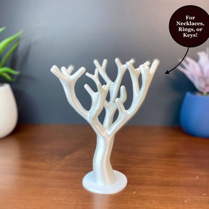 May include: A white, tree-shaped jewelry holder with multiple branches for hanging necklaces, rings, or keys. The holder is made of a smooth material and sits on a round base. The text "For Necklaces, Rings, or Keys!" is in a black circle.