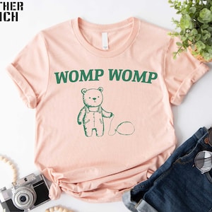 May include: A light peach-colored t-shirt with the words "WOMP WOMP" in green, above a green illustration of a bear holding a balloon. The shirt is folded, with a pair of blue jeans and sunglasses in the background.