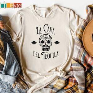 May include: Cream-colored t-shirt featuring a black sugar skull design with the text "La Cava Del Tequila". The shirt is styled with a denim jacket, a brown hat, and a plaid scarf, creating a casual, festive look.