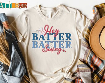 May include: Cream-colored t-shirt with the text "Hey Batter Batter Swing" in blue and red. A baseball graphic is above the text. The shirt is knotted at the waist and is next to a denim jacket and a plaid scarf.