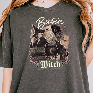 May include: A charcoal gray short-sleeve t-shirt featuring a graphic print of a witch, a cauldron, a black cat, books, and potion bottles. The text "Basic Witch" is printed in white and green. The shirt has a crew neckline and a relaxed fit.