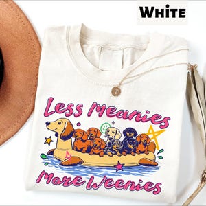 A white t-shirt with a colorful graphic design depicting a group of dogs floating on an inflatable raft, accompanied by the text "Less meanies, more weenies" in pink and purple letters.