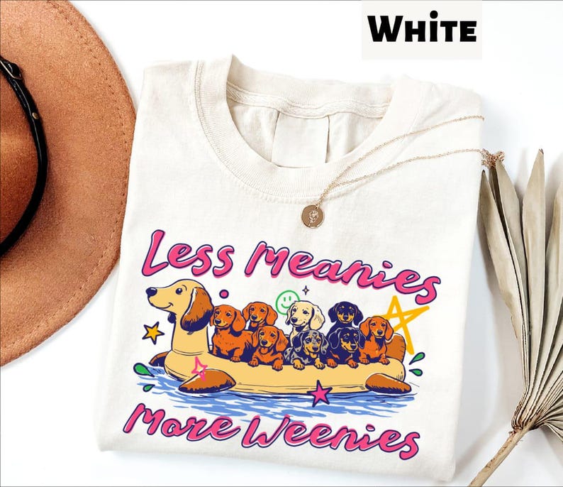 A white t-shirt with a colorful graphic design depicting a group of dogs floating on an inflatable raft, accompanied by the text "Less meanies, more weenies" in pink and purple letters.