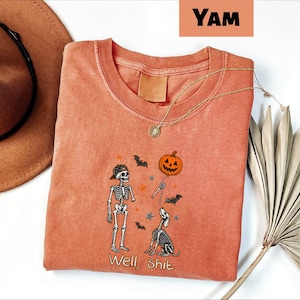 May include: A burnt orange crewneck t-shirt featuring a graphic of a skeleton wearing a baseball cap and a skeleton dog. The design includes a jack-o'-lantern balloon, small bats, and stars, with the text "Well, shit" printed at the bottom.